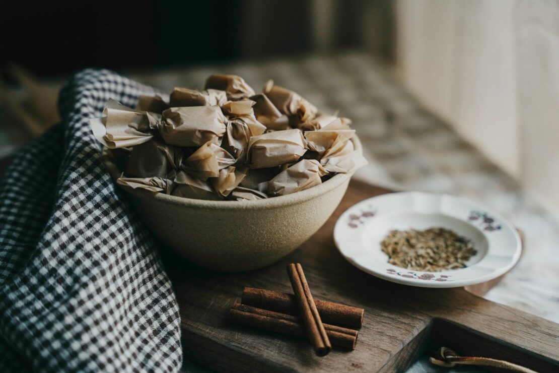 bowl of wrapped caramels with cumin and cinnamon sticks next to it on a table